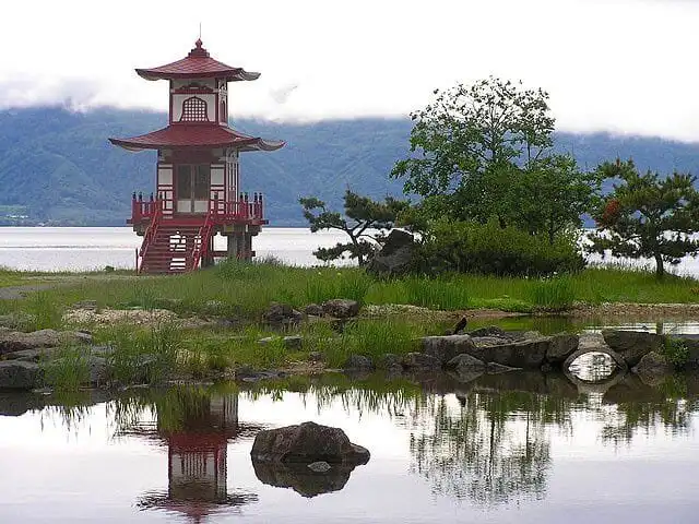 A traditional Japanese-style pavilion by a tranquil lake in Hokkaido, surrounded by lush greenery an.