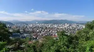 Panoramic view of a Japanese cityscape with lush greenery and distant mountains under a clear blue s.