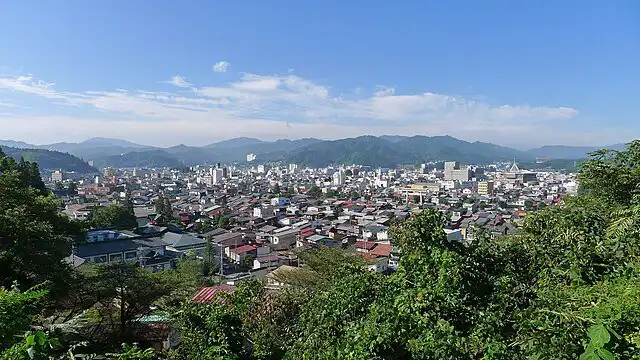 Panoramic view of a Japanese cityscape with lush greenery and distant mountains under a clear blue s.